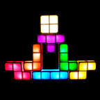 A pyramid-shaped arrangement of glowing light-up block tiles in multiple colors, including yellow, orange, pink, purple, green, and blue, set against a solid black background.